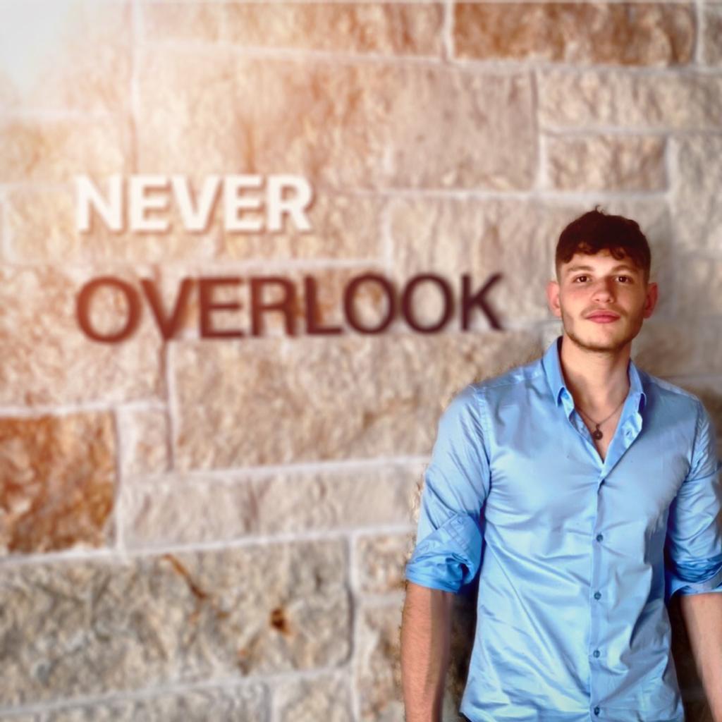 Amit wearing Blue button shirt, standing near a wall with the phrase "never overlook"